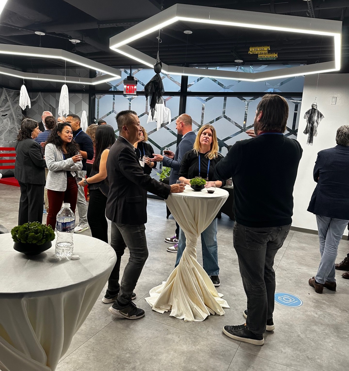 The image shows a group of people socializing at a networking or informal event. They're gathered around cocktail tables, likely in a modern and stylish venue. The space is decorated with a Halloween theme, with hanging bats and other decorations. People are casually talking, enjoying drinks, and engaged in conversation. The atmosphere is relaxed and festive, likely part of a professional gathering or celebration.